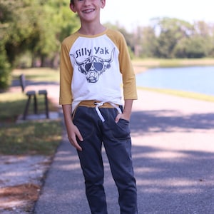 May include: A young person wearing a white and yellow baseball tee shirt with the text "Silly Yak" and a graphic of a cow wearing sunglasses. The person is also wearing dark blue sweatpants and gray sneakers.