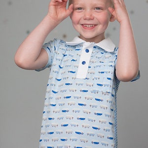 May include: A young boy wearing a light blue short-sleeved polo shirt with a white collar and a pattern of blue and white trucks. The shirt has a blue button closure. The boy is smiling and has blonde hair.