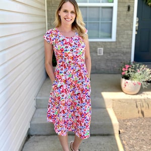 May include: A woman wearing a pink and white floral midi dress with short sleeves and pockets. She is standing on a concrete walkway in front of a house.