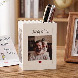 Könnte beinhalten: Ein weißer Stifthalter im Blockstil mit einem Foto von Vater und Kind, den Worten "Daddy & Me" und einem roten Herzen. Zwei Stifte befinden sich im Halter. Ein gerahmtes Foto und eine Vatertagskarte befinden sich ebenfalls auf der Holzoberfläche.