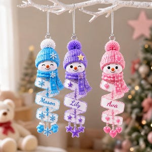 May include: Three hanging snowman ornaments with knitted hats and scarves in blue, purple, and pink. Each snowman features a spiral ribbon tail printed with the names Mason, Lily, and Ava, accented with matching snowflakes, stars, and hearts.