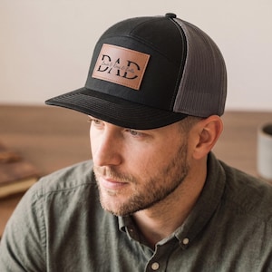 Può includere: Cappellino da camionista nero e grigio con una toppa in pelle con la scritta "DAD". Il cappellino ha una visiera curva e un retro in rete. La toppa include anche i nomi "Louis & Rina & Bella".