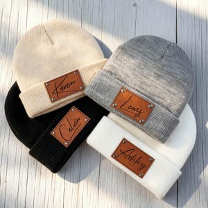 May include: Four knit beanies in cream, gray, black, and white. Each beanie has a brown leather patch with a name: Karen, Lewis, Calvin, and Ashley. The beanies are arranged on a light wood surface.