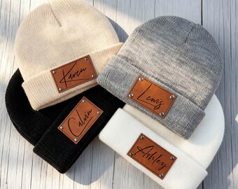 May include: Four knit beanies in cream, gray, black, and white. Each beanie has a brown leather patch with a name: Karen, Lewis, Calvin, and Ashley. The beanies are arranged on a light wood surface.