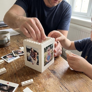 May include: A white Lego block photo frame with family photos. The frame is being assembled with white Lego bricks. A child and an adult are working together on a wooden table. A mug and additional photos are also on the table.