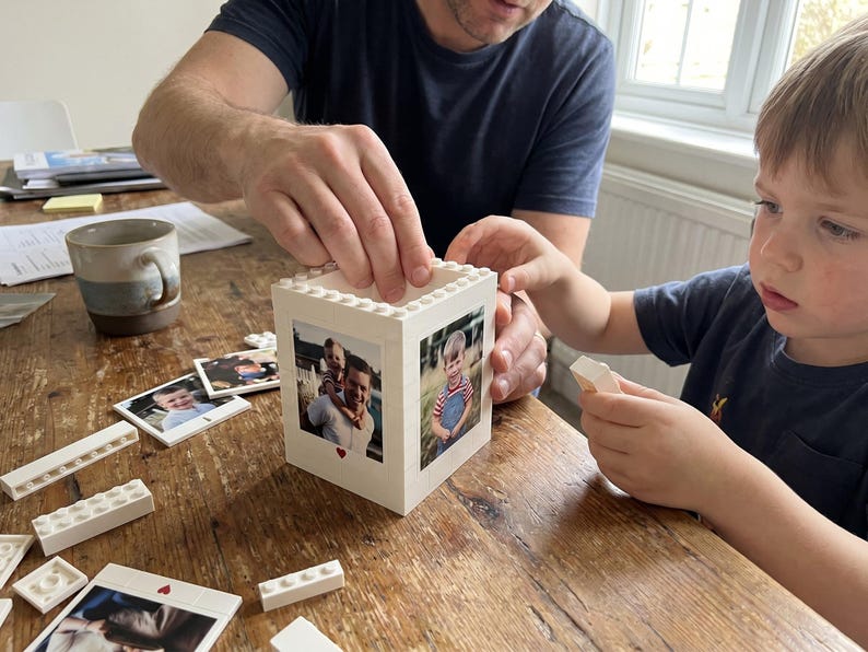 May include: A white Lego block photo frame with family photos. The frame is being assembled with white Lego bricks. A child and an adult are working together on a wooden table. A mug and additional photos are also on the table.