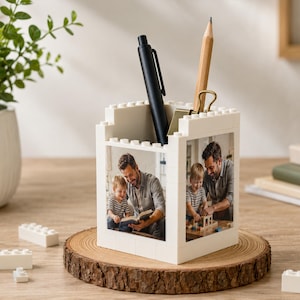 May include: A white desk organizer made of interlocking blocks, featuring a photo of a father and child. A black pen and a pencil with a gold clip are inside. The organizer sits on a wooden slice.