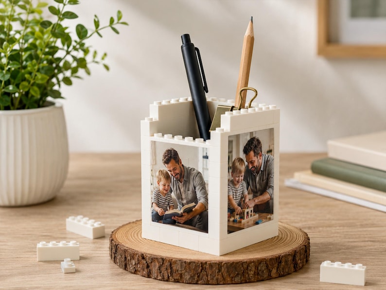 May include: A white desk organizer made of interlocking blocks, featuring a photo of a father and child. A black pen and a pencil with a gold clip are inside. The organizer sits on a wooden slice.