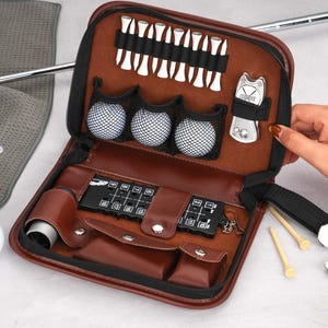 Pode incluir: Um kit de acess&oacute;rios de golfe em couro castanho, aberto para exibir bolas de golfe, tees e uma ferramenta de reparo de divots. O kit inclui um porta-cart&otilde;es e uma pequena bolsa. As bolas de golfe s&atilde;o mantidas em um suporte preto.