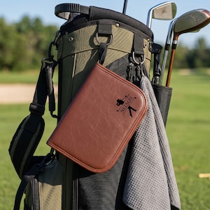 Pode incluir: Um porta-cart&otilde;es de golfe em couro castanho com o nome "Nelson" e uma silhueta de golfista est&aacute; preso a um saco de golfe. O saco &eacute; verde-oliva e preto, com tacos de golfe e uma toalha cinzenta. O fundo &eacute; um campo de golfe verde.