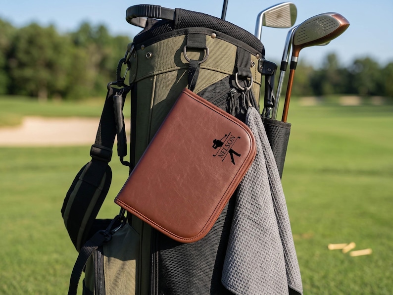 Pode incluir: Um porta-cart&otilde;es de golfe em couro castanho com o nome "Nelson" e uma silhueta de golfista est&aacute; preso a um saco de golfe. O saco &eacute; verde-oliva e preto, com tacos de golfe e uma toalha cinzenta. O fundo &eacute; um campo de golfe verde.