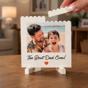 Puede incluir: Un marco de bloques de construcción blancos con una fotografía de un adulto y un niño en la playa. El marco incluye el texto negro The Best Dad Ever! y un pequeño icono de corazón rojo. Una mano coloca un bloque blanco sobre el marco.