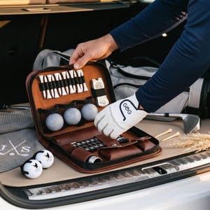 Pode incluir: Um kit de golfe em couro castanho com bolas, tees e acess&oacute;rios. O kit est&aacute; aberto, revelando o seu conte&uacute;do, e est&aacute; a ser segurado por uma pessoa. Uma luva de golfe com as letras "GoG" &eacute; vis&iacute;vel. Equipamento de golfe adicional est&aacute; no fundo.