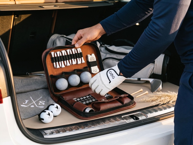 Pode incluir: Um kit de golfe em couro castanho com bolas, tees e acess&oacute;rios. O kit est&aacute; aberto, revelando o seu conte&uacute;do, e est&aacute; a ser segurado por uma pessoa. Uma luva de golfe com as letras "GoG" &eacute; vis&iacute;vel. Equipamento de golfe adicional est&aacute; no fundo.