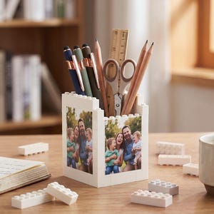 May include: A white brick-style pen holder with a family photo on the sides, filled with pens, pencils, scissors, and a ruler. The desk also has a cup of coffee, a notebook, and some loose bricks.