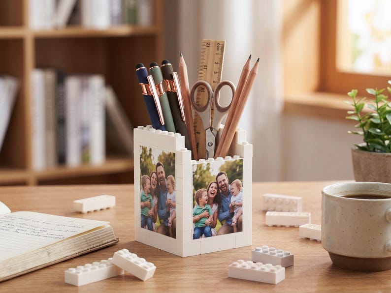 May include: A white brick-style pen holder with a family photo on the sides, filled with pens, pencils, scissors, and a ruler. The desk also has a cup of coffee, a notebook, and some loose bricks.