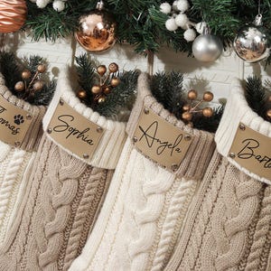 May include: Four cable-knit Christmas stockings in cream and beige tones, each featuring a rectangular tan leather-like name tag with black text reading Thomas, Sophia, Angela, and Barton, with paw prints on the Thomas and Barton tags.