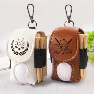Op de afbeelding: Twee golfbalzakjes, één witte en één bruine, elk met een golfbal, tees en een zilveren karabijnhaak. Het witte zakje heeft de initialen "BXS" en een golf-thema embleem. Het bruine zakje heeft de naam "Xavier".