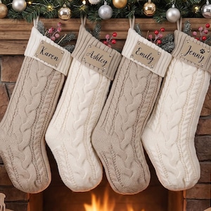 Puede incluir: Cuatro calcetines navideños de punto trenzado en colores beige y crema alternos, cada uno con un puño acanalado y una etiqueta de nombre de cuero sintético personalizada. Los nombres Karen, Ashley, Emily y Jenny están impresos, con una huella.