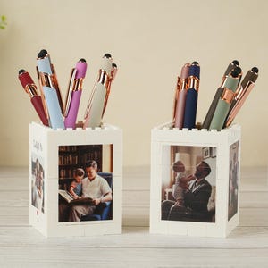 May include: Two white block-style pen holders with family photos. Each holds several pens in various colors, including burgundy, blue, and green, with gold-colored accents. The pen holders are on a light-colored wooden surface.