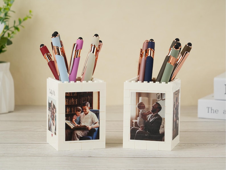 May include: Two white block-style pen holders with family photos. Each holds several pens in various colors, including burgundy, blue, and green, with gold-colored accents. The pen holders are on a light-colored wooden surface.