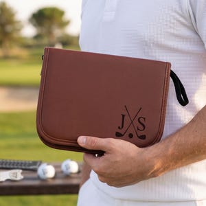 Pode incluir: Uma bolsa de acess&oacute;rios de golfe de couro marrom com uma al&ccedil;a preta e as iniciais "JXS" com tacos de golfe cruzados e uma bola de golfe. Duas bolas de golfe e um marcador de tee est&atilde;o em uma mesa de madeira no fundo.