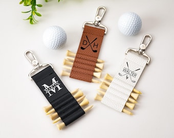 May include: Three personalized golf tee holders with silver clasps, each holding several wooden tees. One is black with the name "MARTIN" in white, another is brown with initials, and the third is white with the name "WALTER". Two golf balls are also present.