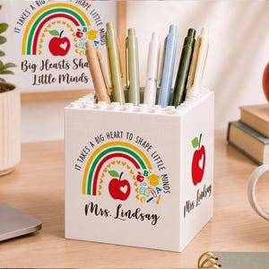 May include: A white square pencil holder constructed from building blocks, featuring a colorful rainbow, a red apple with a heart, and the text "It takes a big heart to shape little minds" and "Mrs. Lindsay" printed on the side, filled with pens.