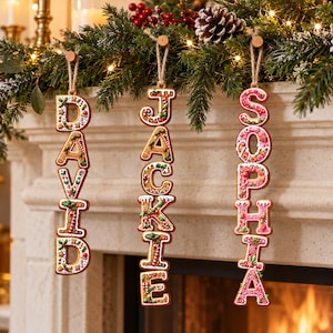 May include: Three vertical hanging ornaments shaped like gingerbread cookies, each featuring a name spelled out in capital letters: DAVID, JACKIE, and SOPHIA. The letters are decorated with white icing, red and green accents, and candy-like details.