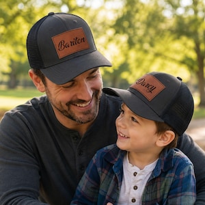 May include: Two charcoal gray trucker hats with black mesh backs and curved brims. Each hat features a rectangular brown faux leather patch on the front. The larger hat has the name Bariton printed in black, and the smaller hat has Darcy printed.