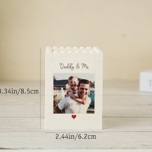 May include: A white block-style photo holder with a picture of a man and child. The text "Daddy & Me" is above the photo, and a red heart is below. The holder is 3.34 inches tall and 2.44 inches wide.