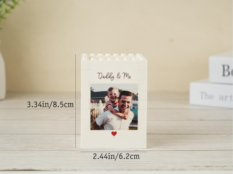 May include: A white block-style photo holder with a picture of a man and child. The text "Daddy & Me" is above the photo, and a red heart is below. The holder is 3.34 inches tall and 2.44 inches wide.