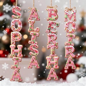 May include: Four vertical hanging ornaments shaped like gingerbread letters spelling the names Sophia, Alisha, Bertha, and Moira. Each letter features pink icing, white decorative accents, and small green and red holiday-themed details.