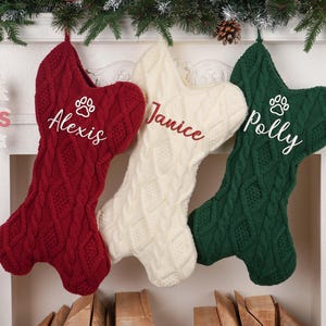 May include: Three cable-knit Christmas stockings in red, cream, and green hang from a mantel. Each stocking is personalized with a name and a paw print. Festive holiday decor.