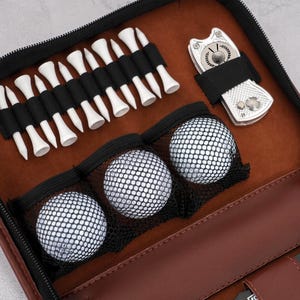 Pode incluir: Uma bolsa de acess&oacute;rios de golfe em couro castanho com z&iacute;per preto, contendo tees de golfe brancos, tr&ecirc;s bolas de golfe e uma ferramenta de reparo de divot prateada. O interior &eacute; de cor castanho claro.