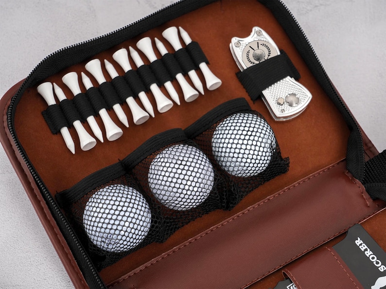 Pode incluir: Uma bolsa de acess&oacute;rios de golfe em couro castanho com z&iacute;per preto, contendo tees de golfe brancos, tr&ecirc;s bolas de golfe e uma ferramenta de reparo de divot prateada. O interior &eacute; de cor castanho claro.