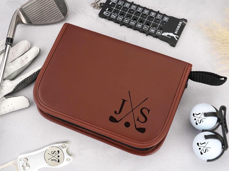 Pode incluir: Uma bolsa de acess&oacute;rios de golfe em couro castanho com z&iacute;per e al&ccedil;a pretos. A bolsa &eacute; personalizada com as iniciais "J S" e tacos de golfe cruzados. Bolas de golfe, um taco de golfe e um marcador de pontua&ccedil;&atilde;o tamb&eacute;m s&atilde;o vis&iacute;veis.
