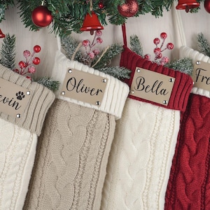 Peut inclure: Quatre bas de Noël en tricot torsadé crème, beige et rouge, chacun avec une étiquette en cuir personnalisée. Les étiquettes affichent les noms : Kevin, Oliver, Bella et Freda. Des décorations festives ornent l'arrière-plan.
