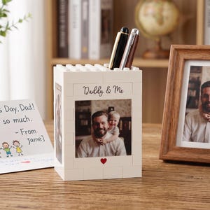 May include: A white block-style pen holder with a photo of a father and child, the words "Daddy & Me", and a red heart. Two pens are in the holder. A framed photo and a Father's Day card are also on the wooden surface.
