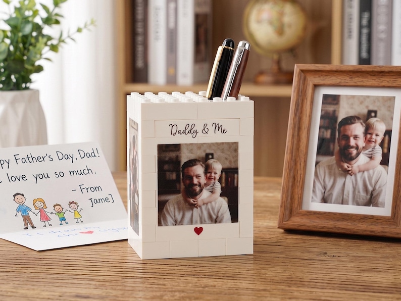 May include: A white block-style pen holder with a photo of a father and child, the words "Daddy & Me", and a red heart. Two pens are in the holder. A framed photo and a Father's Day card are also on the wooden surface.