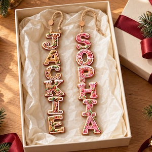 May include: Two vertical hanging ornaments shaped like gingerbread cookies, spelling the names JACKIE and SOPHIA. The letters feature white icing details, red and green accents, and are suspended by tan twine loops inside a white gift box.