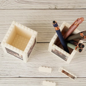 May include: Two white, brick-style desk organizers with a printed design. One holds several pens in shades of blue, pink, and green with rose gold accents. The other is empty. The organizers sit on a light wood surface.