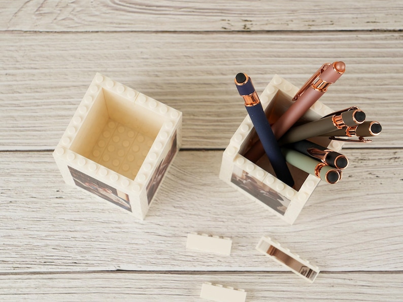 May include: Two white, brick-style desk organizers with a printed design. One holds several pens in shades of blue, pink, and green with rose gold accents. The other is empty. The organizers sit on a light wood surface.
