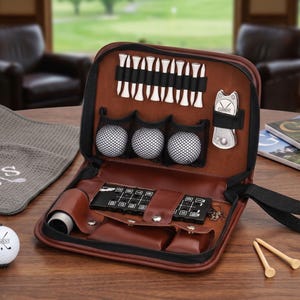 Pode incluir: Um kit de golfe em couro castanho com z&iacute;per preto, contendo bolas de golfe, tees, uma ferramenta de reparo de divots e um porta-cart&otilde;es. Duas bolas de golfe e uma toalha cinza com bordado de tacos de golfe tamb&eacute;m s&atilde;o vis&iacute;veis.