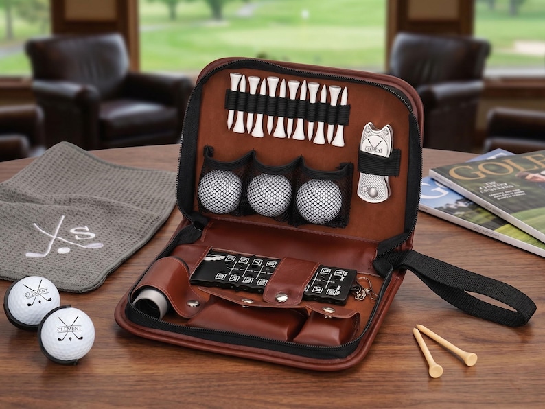 Pode incluir: Um kit de golfe em couro castanho com z&iacute;per preto, contendo bolas de golfe, tees, uma ferramenta de reparo de divots e um porta-cart&otilde;es. Duas bolas de golfe e uma toalha cinza com bordado de tacos de golfe tamb&eacute;m s&atilde;o vis&iacute;veis.