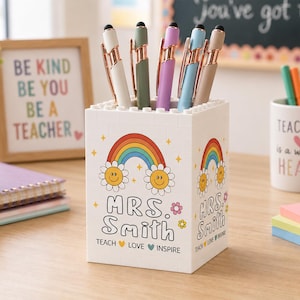 May include: A white square pen holder constructed from building blocks, featuring a rainbow and smiley face flower graphic. The text reads "MRS. SMITH" and "TEACH LOVE INSPIRE". It holds five pens with rose gold clips and stylus tips.