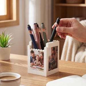 May include: A collection of pens in a white block-style pen holder with family photos. The pens are various colors, including navy, pink, and green, with gold accents. A hand is reaching for a black pen with a gold tip.