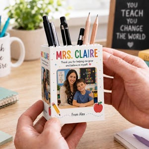Puede incluir: Un portalápices cuadrado blanco con el texto "MRS. CLAIRE" en colores arcoíris, una fotografía impresa de un docente y un estudiante, la frase "Thank you for helping me grow and believe in myself" y "From Alex" con gráficos de lápiz y manzana.