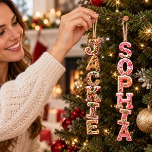 May include: Two vertical Christmas tree ornaments shaped like gingerbread cookies, featuring the names JACKIE and SOPHIA in white icing. The letters are decorated with colorful sprinkles and red and green accents, each hanging from a rustic twine loop.