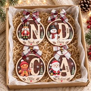 May include: Four circular wooden Christmas ornaments in a gift box. Each features a large letter, the year 2026, a holiday character, and a plaid bow. Characters include a nutcracker, snowman, gingerbread person, and Santa. Names Noah, Daisy, Chloe, and Andrew are engraved at the bottom of each ornament.